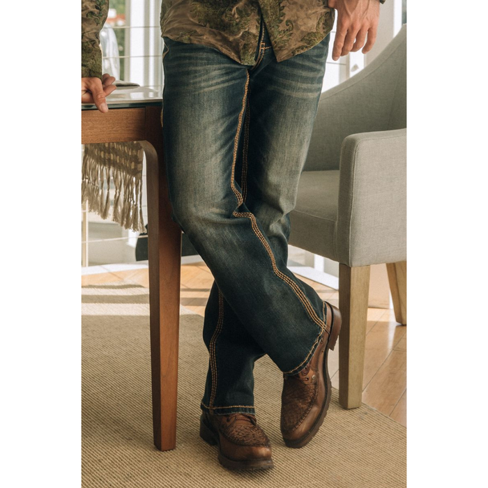 Indoor lifestyle image of model wearing Platini Holt Dark Stone Slim Boot Cut Jeans leaning against a table.